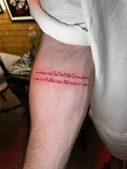 Two lines of Pali verse in Thai script tattooed in red ink on the forearm